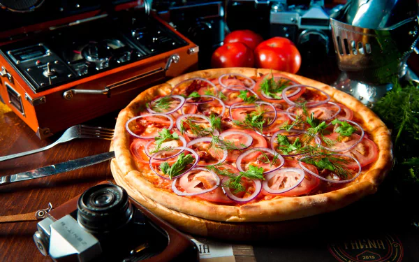 HD desktop wallpaper still life: pepperoni and red onion pizza with herbs beside a Zenit camera, tomatoes and kitchen tools on a rustic table.
