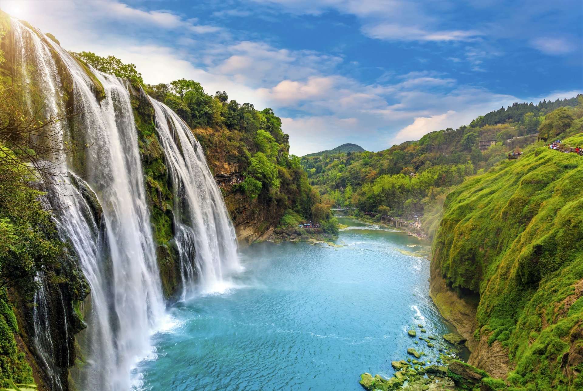 4K Ultra HD desktop wallpaper featuring a vibrant waterfall cascading into a serene river surrounded by lush green hills under a bright blue sky.