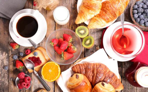 8K Ultra HD PC desktop wallpaper of a breakfast spread: croissants and viennoiserie, strawberries, blueberries, kiwi and orange, toast with jam, cup of coffee and a tea cup on a wooden table.