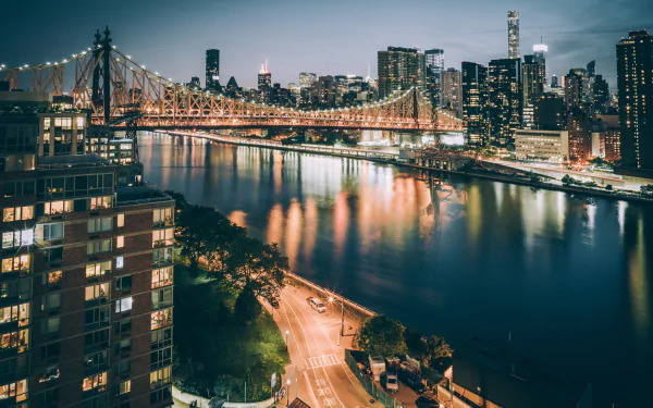 Night view of the Queensboro Bridge spanning the East River, with illuminated skyscrapers and city buildings in New York, USA, reflecting on the water.