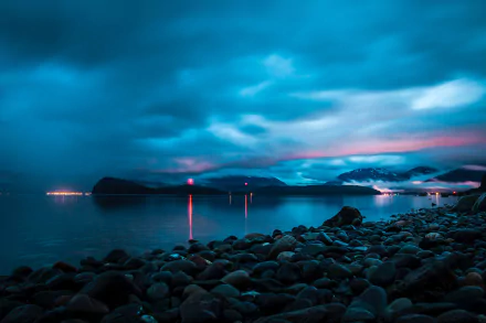 HD desktop wallpaper of an Alaskan coastline at dusk, with a rocky shore, calm waters, distant lights, and a dramatic, cloudy sky.