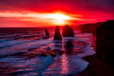 Sunset over the ocean at The Twelve Apostles along the coastline of Victoria, Australia, with vibrant red and orange hues illuminating the sky and water.