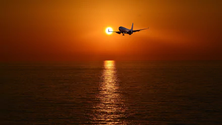 Passenger plane flying over the ocean at sunset, with the sun near the horizon casting a warm orange glow, captured in an HD desktop wallpaper background.