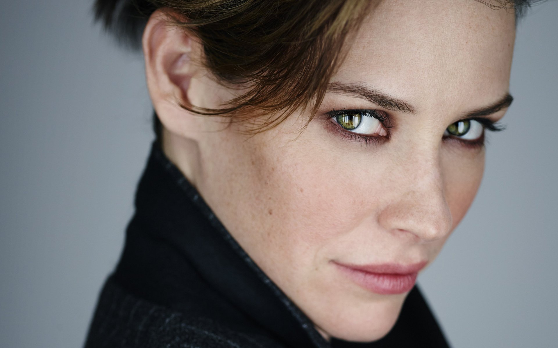 Close-up portrait of a woman with short brown hair and striking green eyes, wearing a dark coat; 2K Quad HD PC desktop wallpaper/background.
