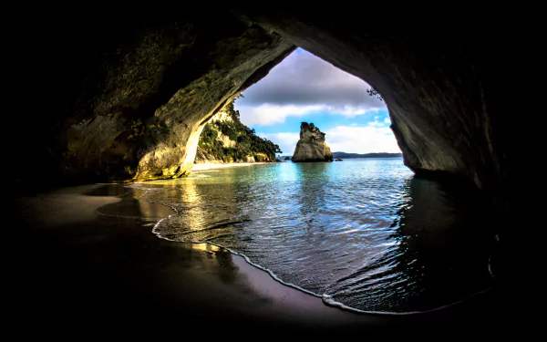 4K Ultra HD wallpaper showing a serene beach view from inside a natural cave, with calm water and a rocky island framed by the cave’s arch.