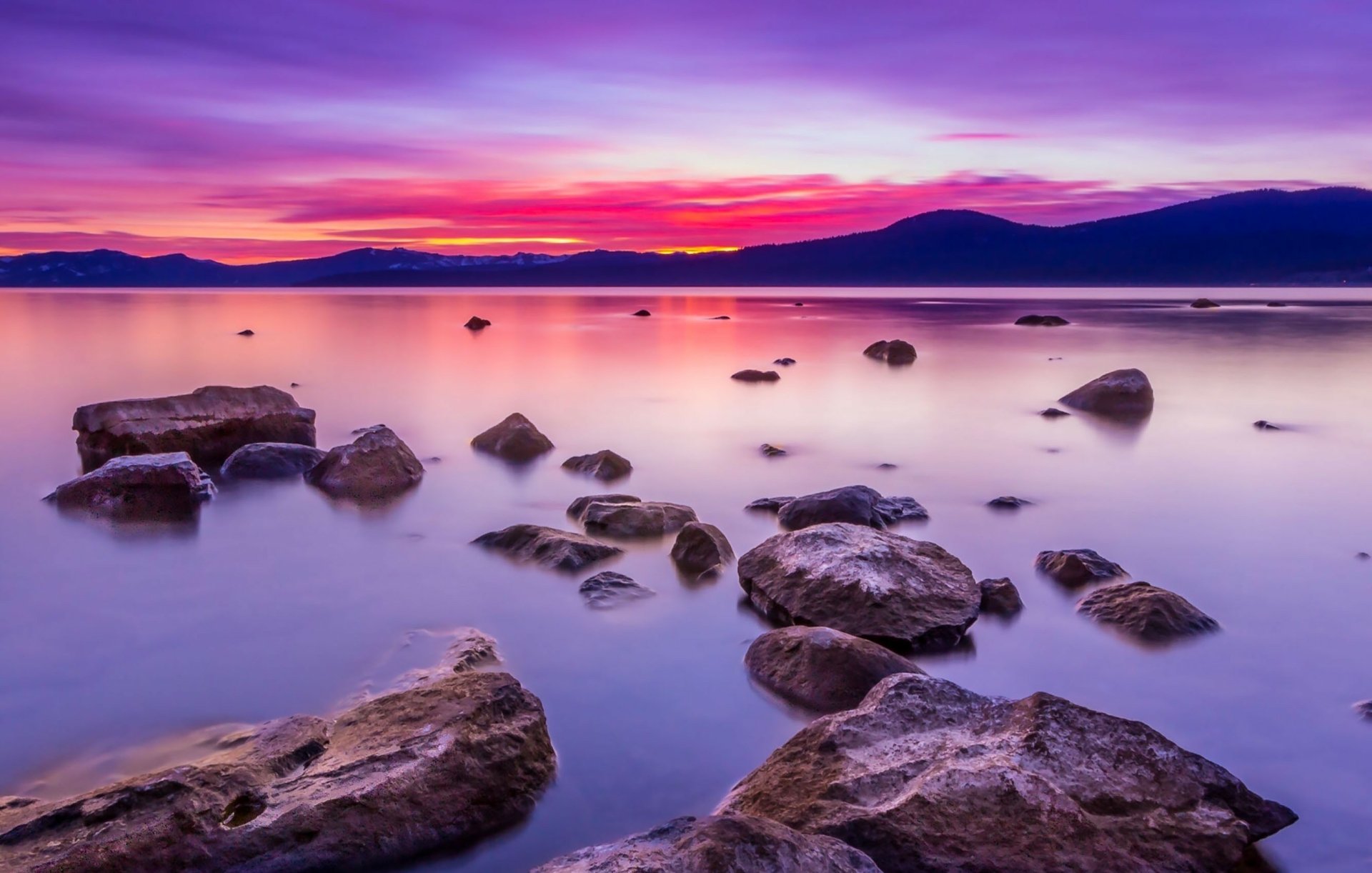 A serene ocean view during sunset with rocks scattered in the calm water, under a vibrant purple and pink sky. HD nature-themed desktop wallpaper and background.