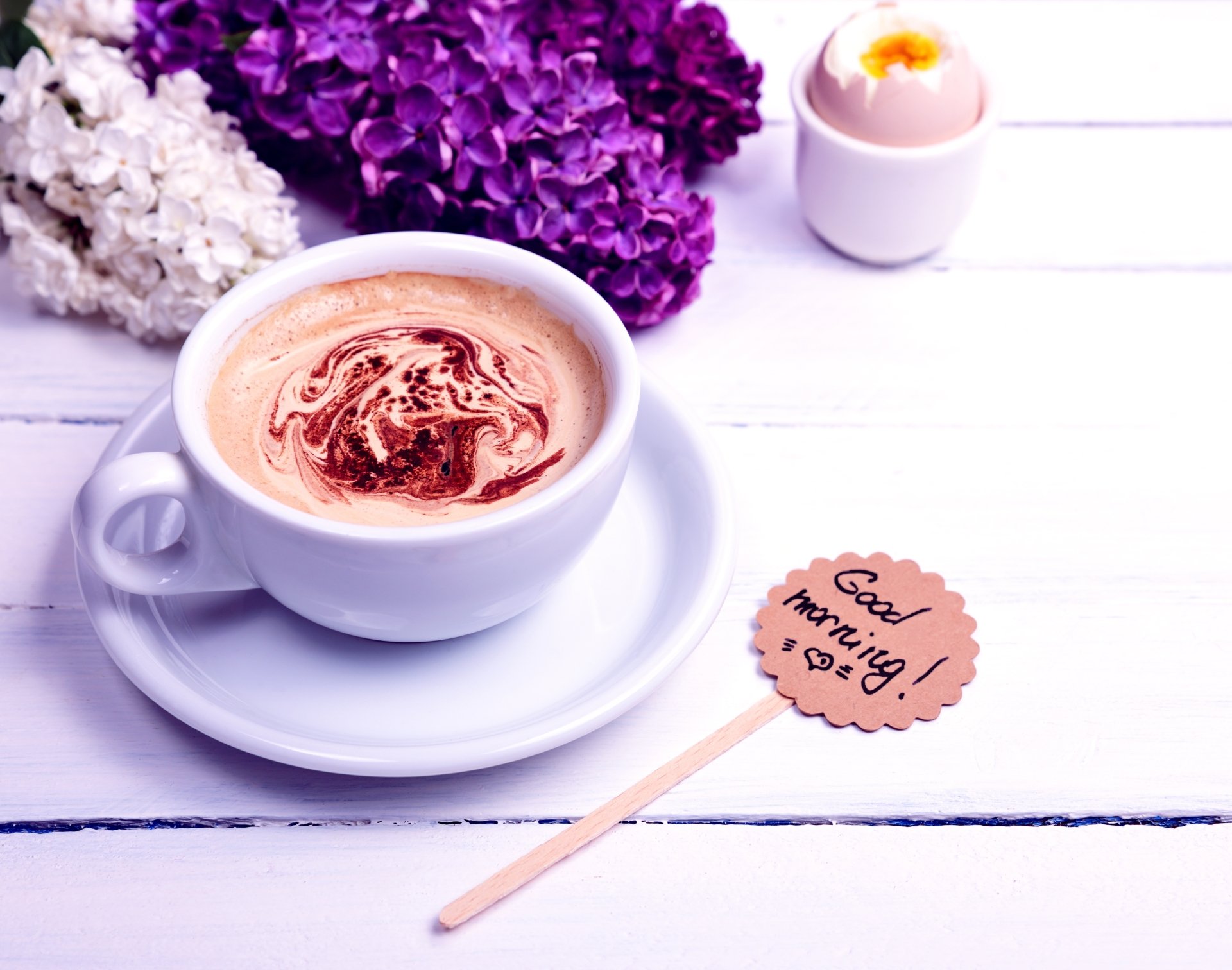 A close-up of a coffee cup with frothy swirls on a white wooden table, accompanied by purple and white flowers and a Good morning! sign, in 4K Ultra HD desktop wallpaper quality.
