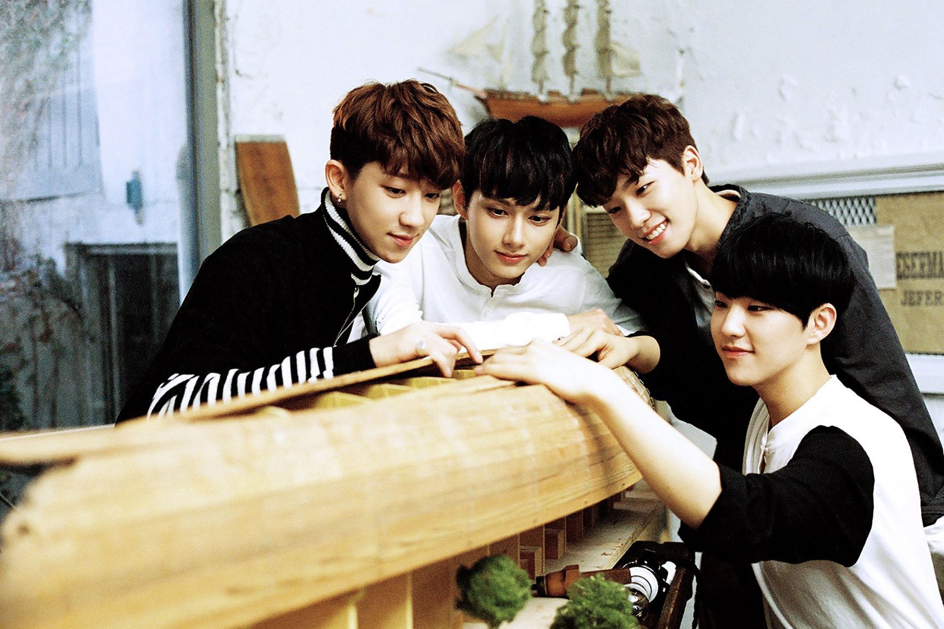 HD PC desktop wallpaper of four smiling band members leaning over a wooden model in a bright studio, casual outfits and warm, music-themed vibe