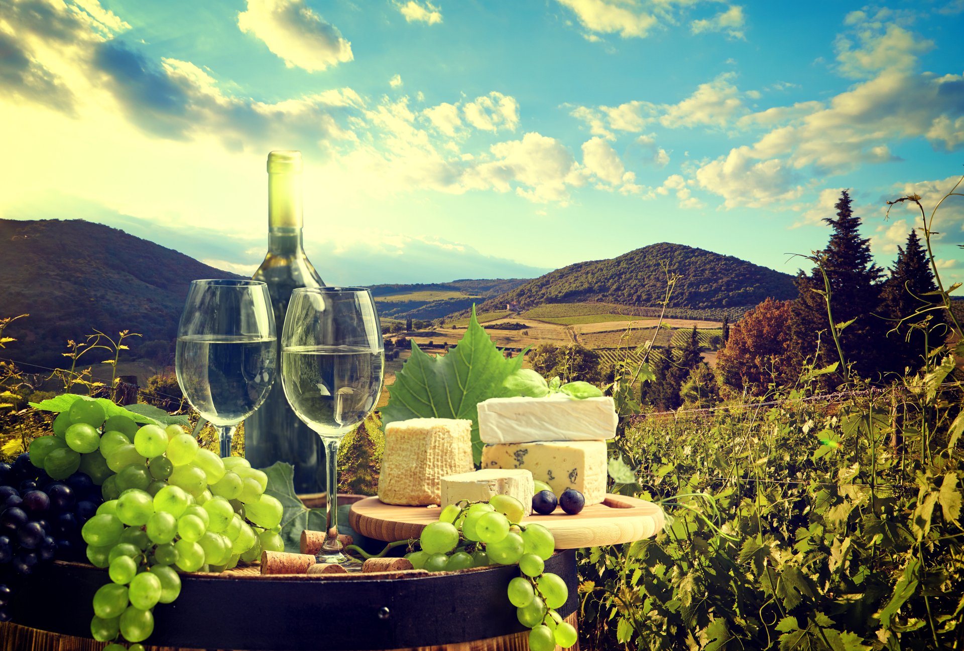 4K Ultra HD still life of wine, grapes, and cheese set against a vibrant vineyard landscape under a partly cloudy sky.