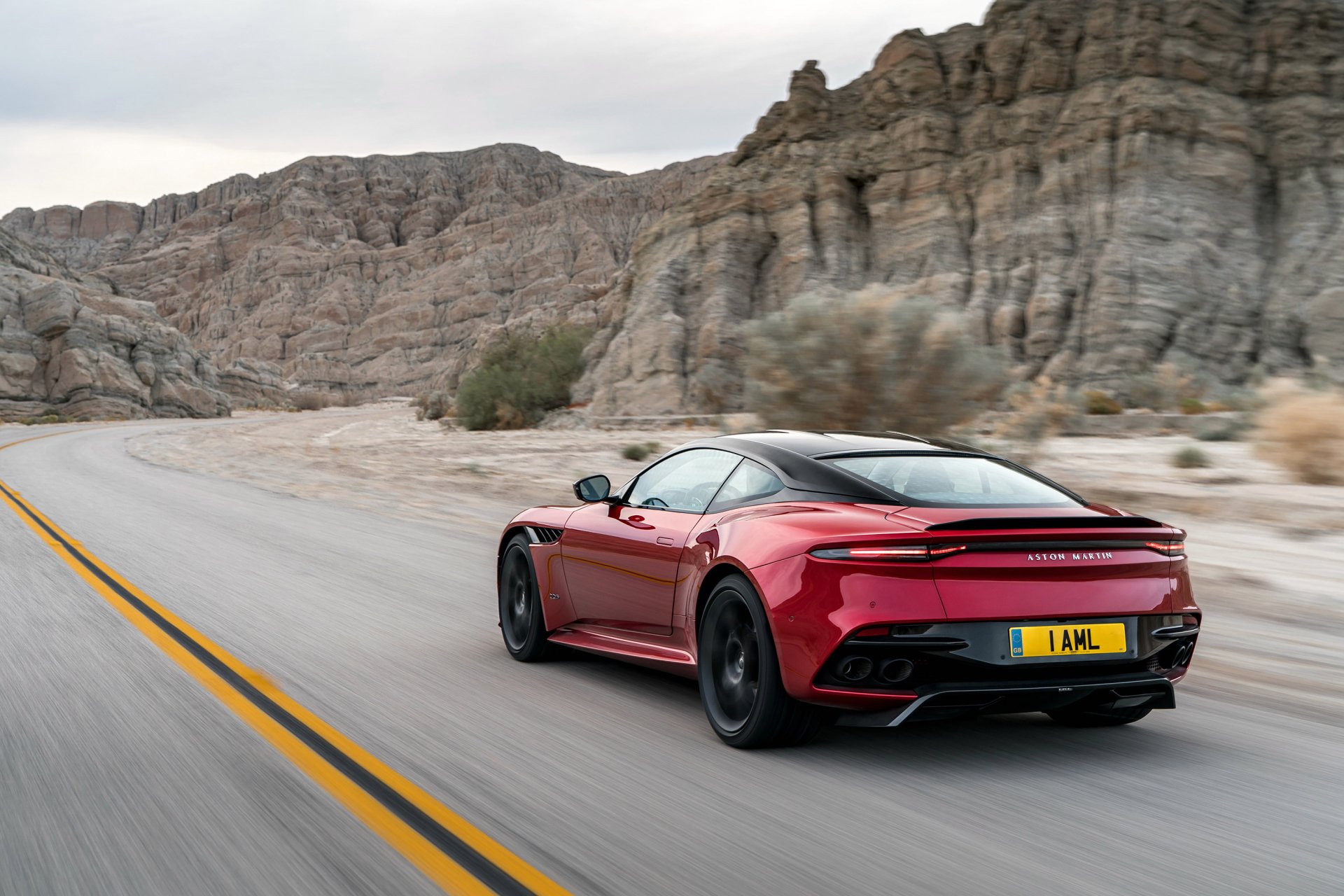 Red Aston Martin DBS Superleggera supercar speeding on a desert road, set against rocky cliffs, captured in a high-definition PC desktop wallpaper.