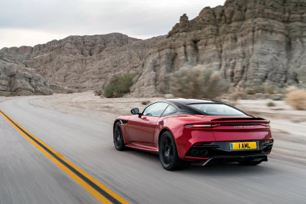 Red Aston Martin DBS Superleggera supercar speeding on a desert road, set against rocky cliffs, captured in a high-definition PC desktop wallpaper.