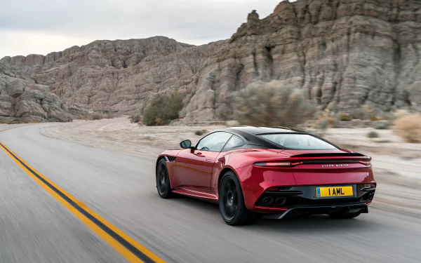 Red Aston Martin DBS Superleggera supercar speeding on a desert road, set against rocky cliffs, captured in a high-definition PC desktop wallpaper.