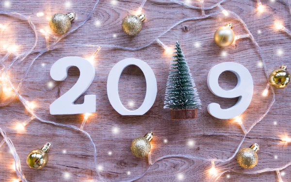 A 4K Ultra HD New Year 2019 desktop wallpaper featuring white numbers, a small Christmas tree, golden baubles, and festive string lights on a wooden background.