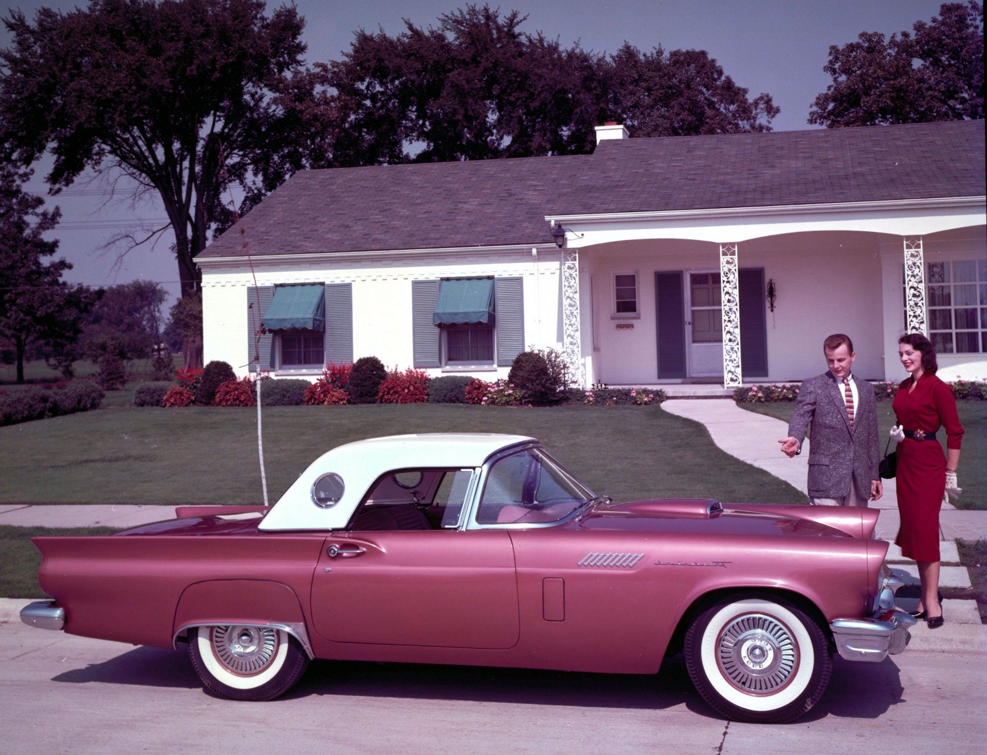 HD PC desktop wallpaper of a vintage pink Ford Thunderbird parked before a midcentury suburban house, with a well-dressed man and woman standing beside the car.