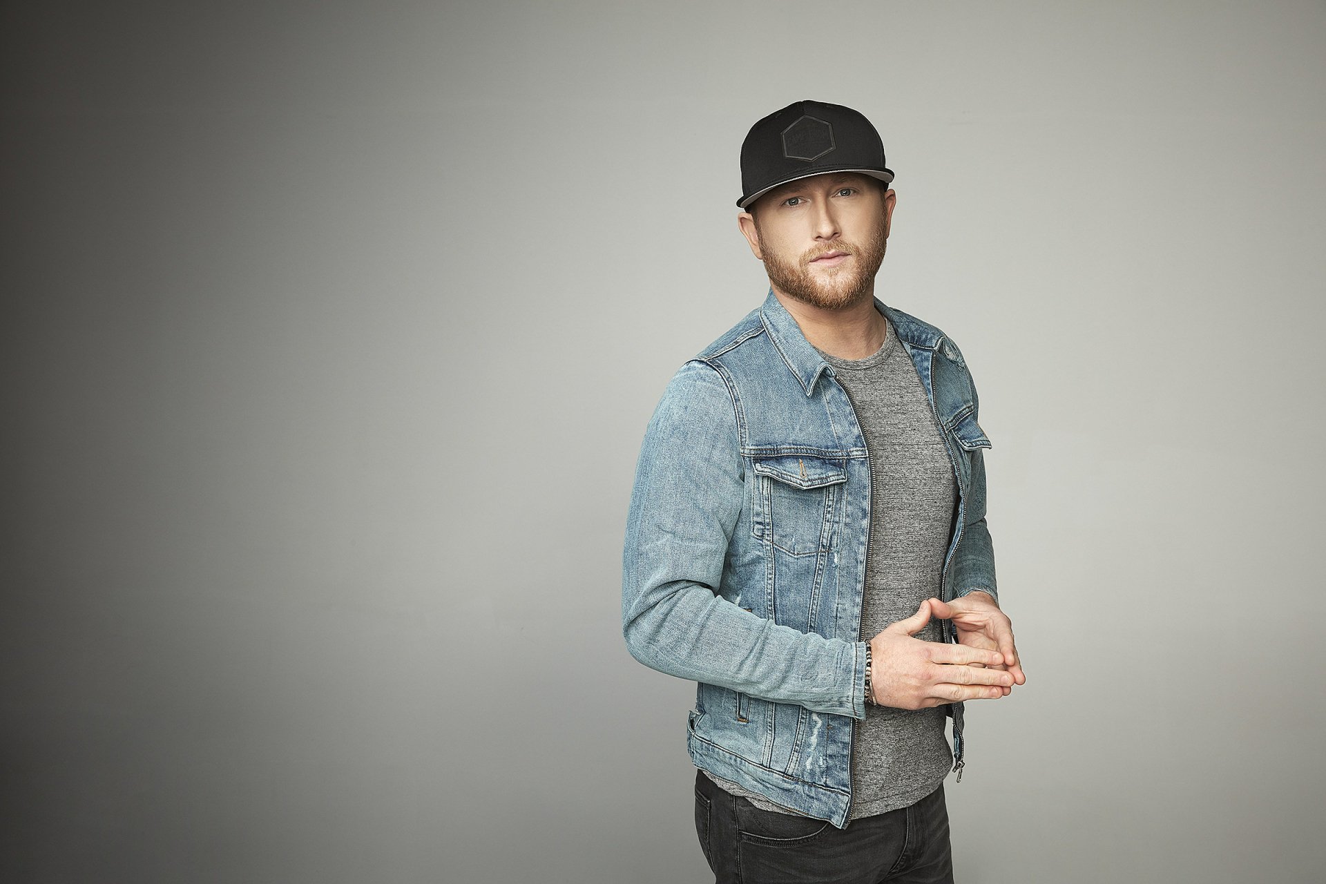 A stylish HD desktop wallpaper featuring country music artist Cole Swindell, captured in a modern look with a denim jacket and a cap against a minimalistic background.