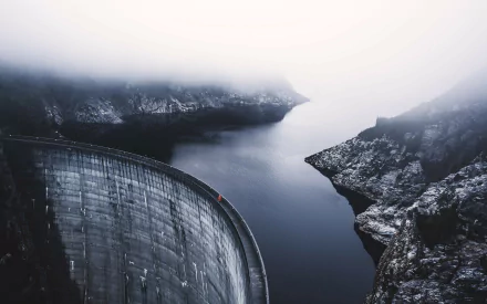 Curved concrete dam spilling into a fog-shrouded river canyon, a moody man-made structure framed by rocky cliffs — 2K Quad HD PC desktop wallpaper/background.