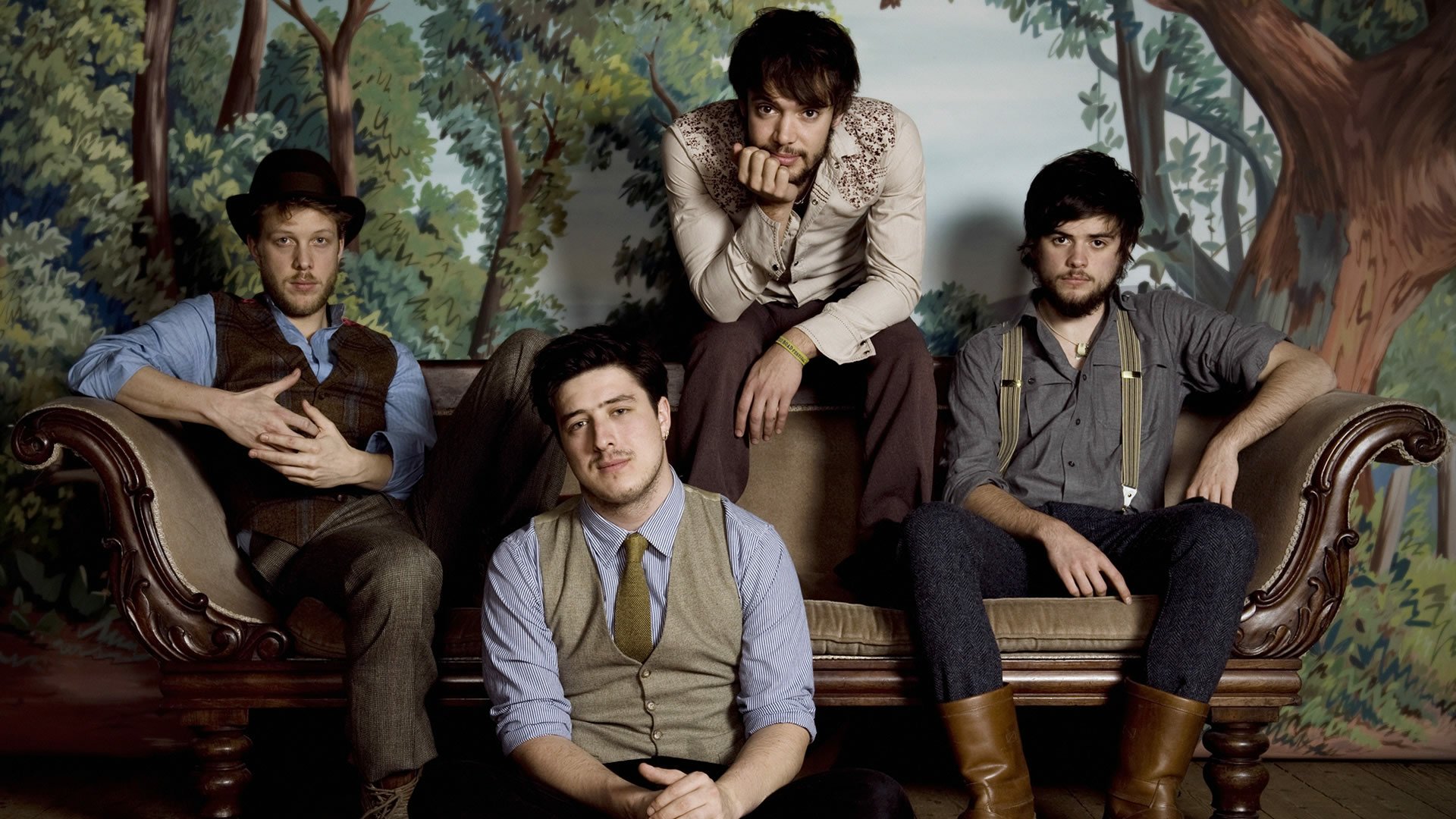 A stylish HD wallpaper featuring Mumford & Sons, showcasing the band members posed comfortably on a vintage sofa against a nature-themed backdrop.