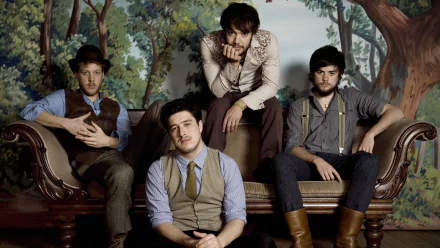 A stylish HD wallpaper featuring Mumford & Sons, showcasing the band members posed comfortably on a vintage sofa against a nature-themed backdrop.