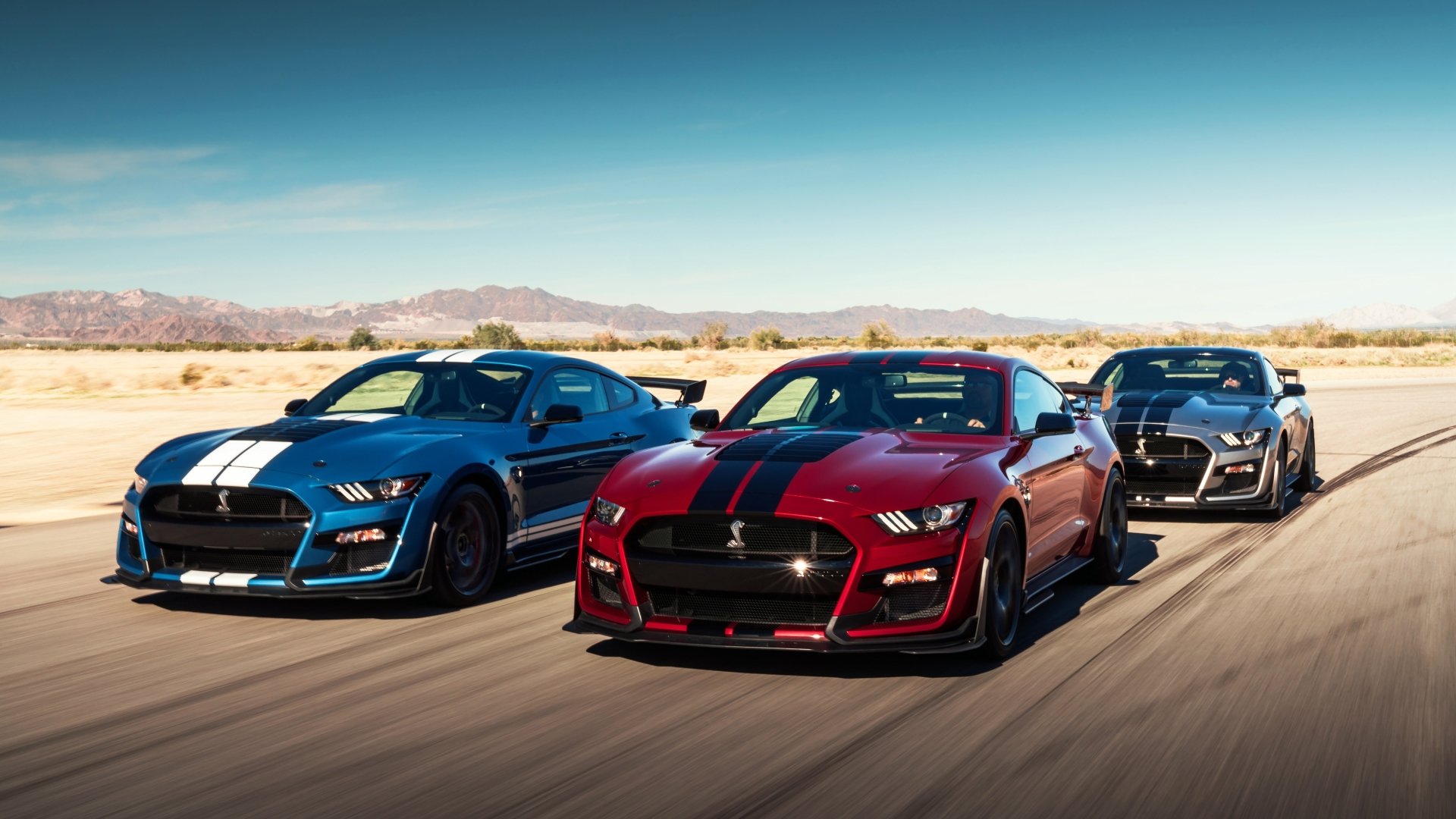 A trio of Ford Mustang Shelby GT500 muscle cars in silver, red, and black drive on a desert road under a clear blue sky, captured in 4K Ultra HD.