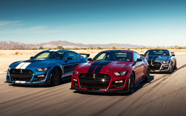 A trio of Ford Mustang Shelby GT500 muscle cars in silver, red, and black drive on a desert road under a clear blue sky, captured in 4K Ultra HD.