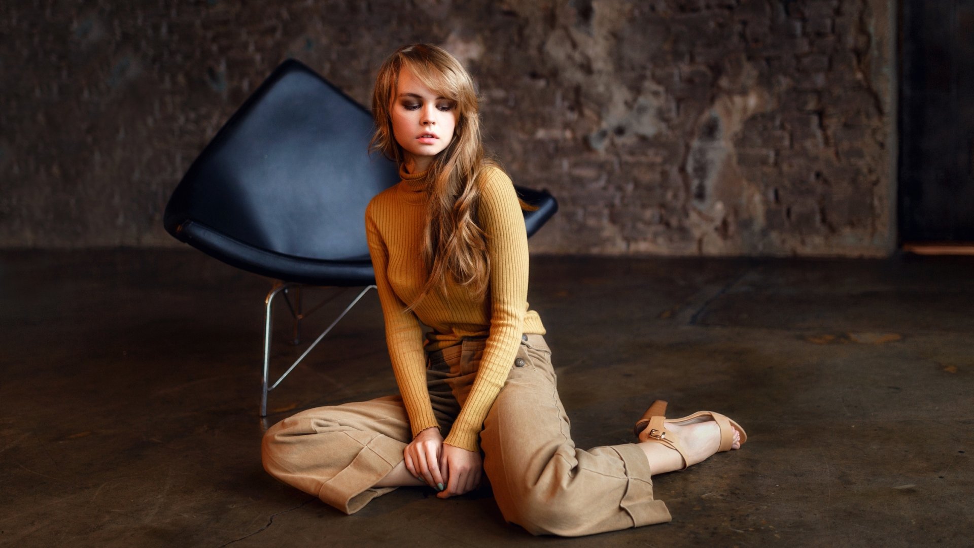 Moody 2K Quad HD desktop wallpaper: a blonde Russian model with long hair in a mustard top and khaki pants, seated on the floor beside a modern chair.