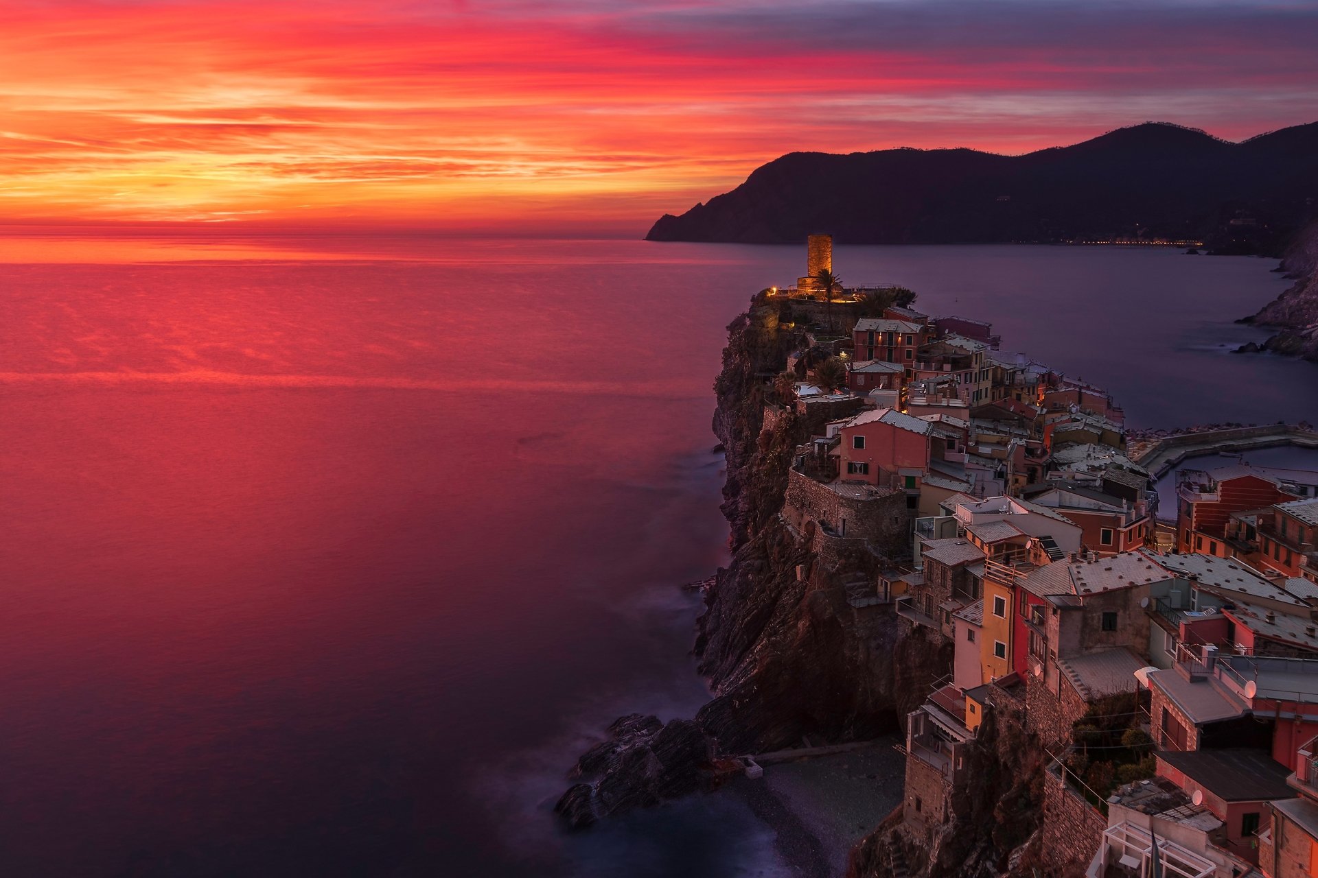 Sunset over Vernazza village in Cinque Terre, with the vibrant horizon casting warm hues over the coastal cliffs and man-made buildings, captured in HD.