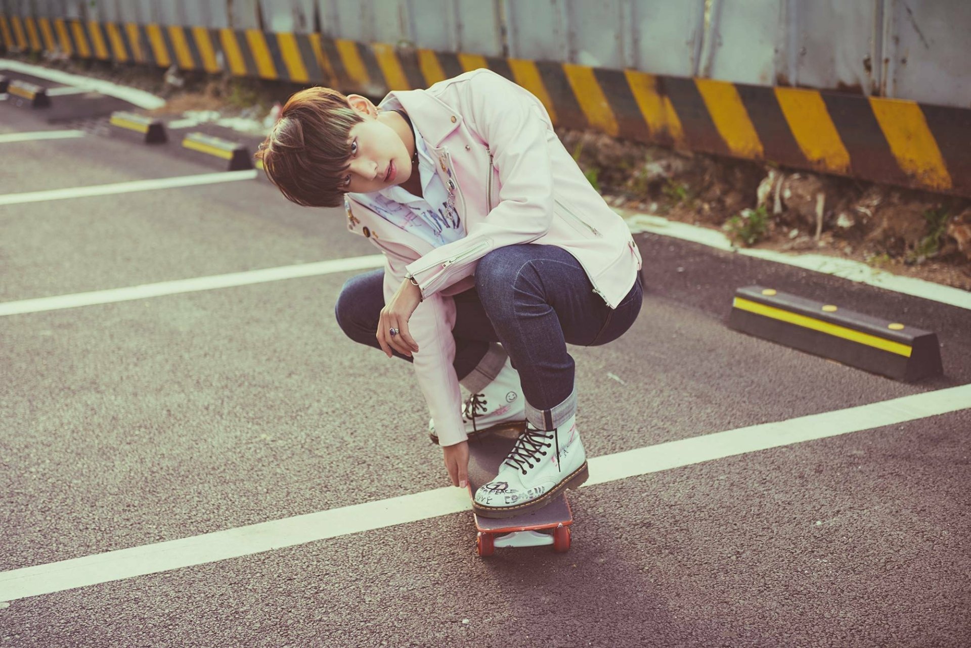 HD desktop wallpaper featuring a member of the K-pop group BTS in casual attire skateboarding in a parking lot with yellow-black striped barriers.