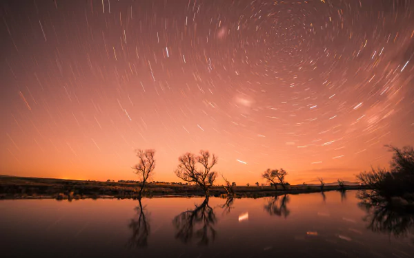 4K Ultra HD wallpaper of star trails swirling above silhouetted trees reflected in calm water during a vibrant sunset in a natural landscape.