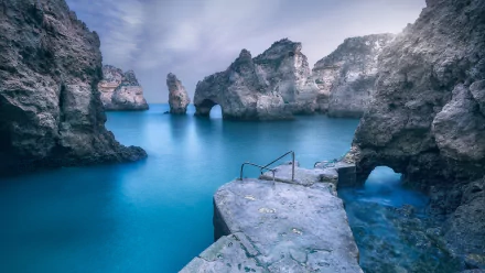 HD desktop wallpaper showcasing Portugal's natural coastal beauty with rugged cliffs, sea arches, and calm turquoise waters under a soft, cloudy sky.