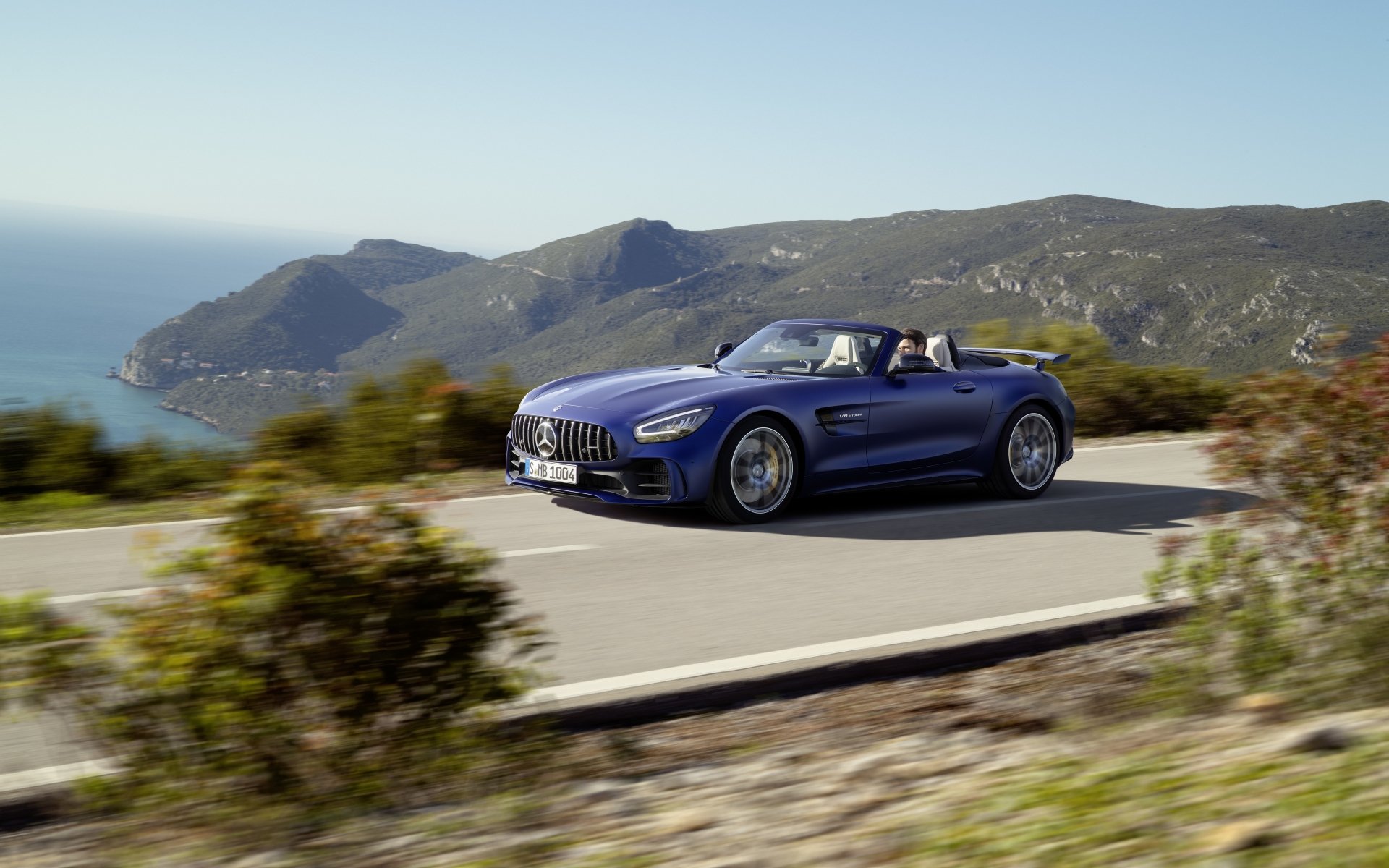 A blue Mercedes-AMG GT R supercar drives along a coastal road with mountains and sea in the background, captured in 4K Ultra HD as a PC desktop wallpaper.