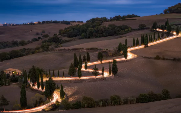 Time-lapse photography captures a winding road illuminated at dusk through the rolling hills of Tuscany, Italy, creating a dynamic HD desktop wallpaper landscape.