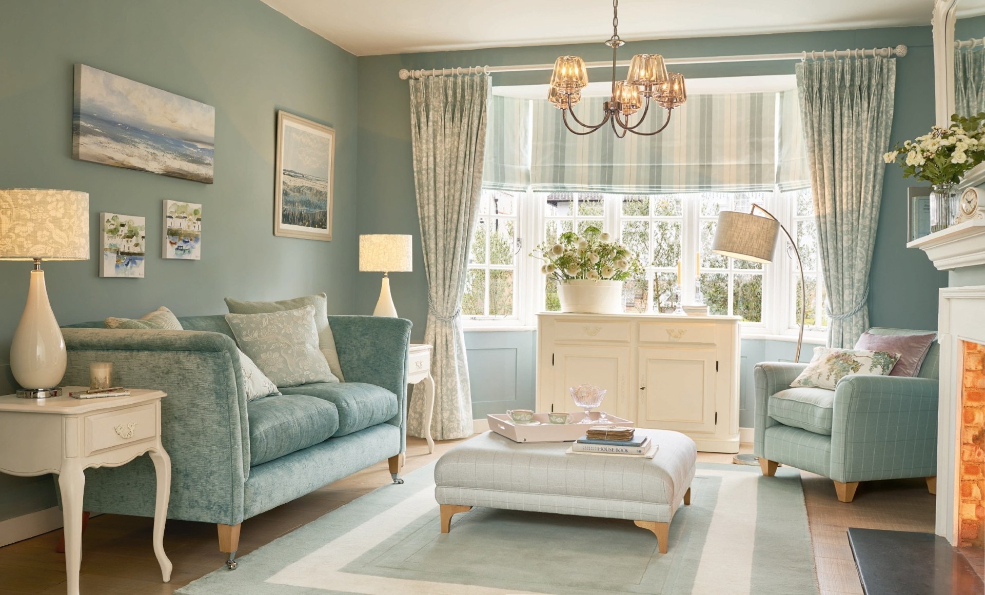 HD wallpaper of a cozy living room with muted blue walls, elegant furniture, soft lighting, and large windows adorned with curtains. The room features a comfortable loveseat, armchair, and a central ottoman.