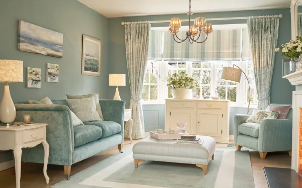 HD wallpaper of a cozy living room with muted blue walls, elegant furniture, soft lighting, and large windows adorned with curtains. The room features a comfortable loveseat, armchair, and a central ottoman.