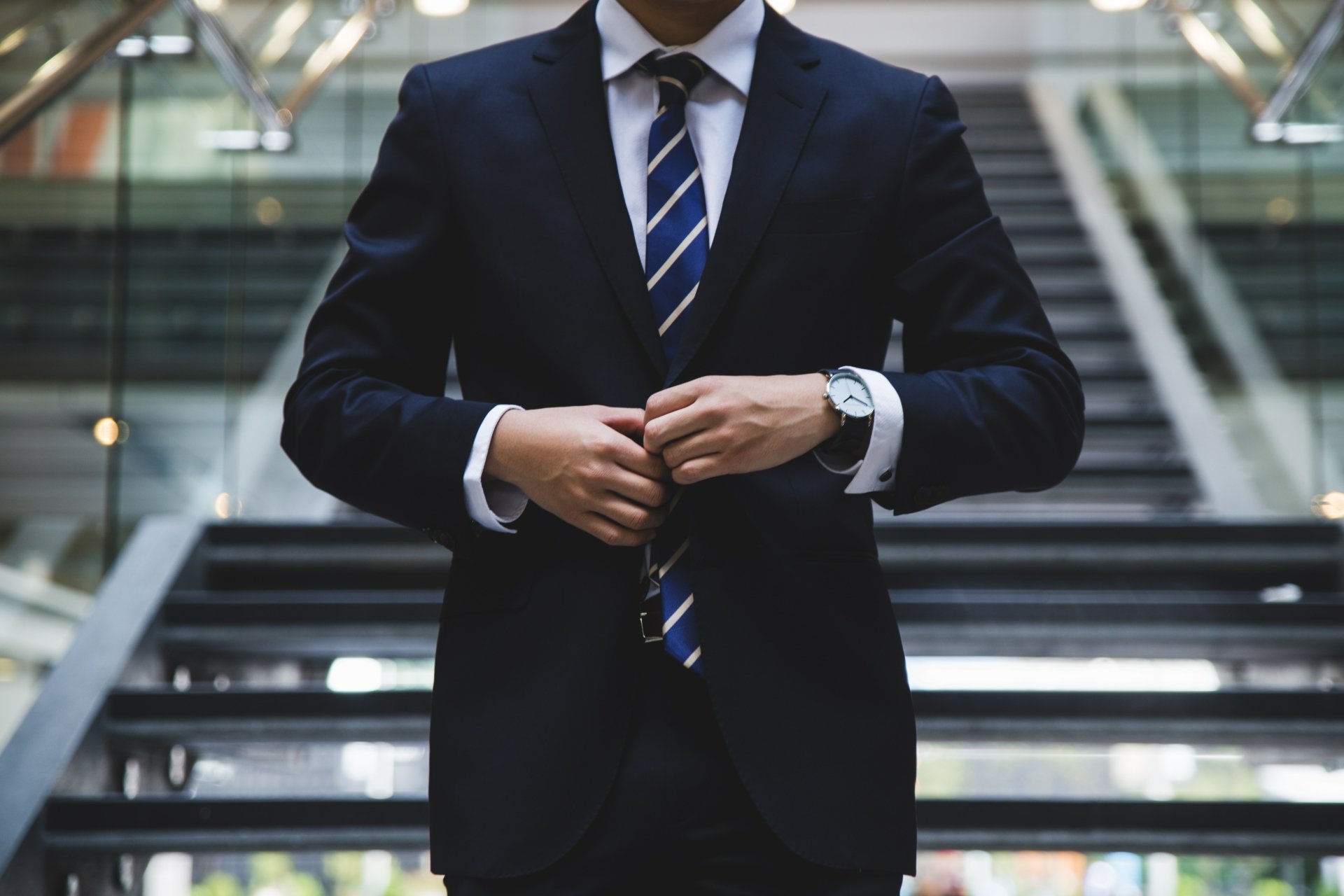 Man in navy suit and striped tie adjusts jacket on modern glass staircase — fashion photography of men. 4K Ultra HD PC desktop wallpaper/background.
